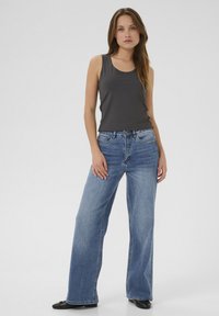 Donna in piedi con pantaloni jeans larghi sbiaditi blu, canotta grigia scura senza maniche e ballerine nere su uno sfondo chiaro e semplice.
