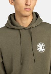 Sudadera de color verde oliva hecha de una mezcla de algodón, con un bolsillo tipo canguro en el frente y un logotipo circular blanco que presenta elementos naturales y texto.