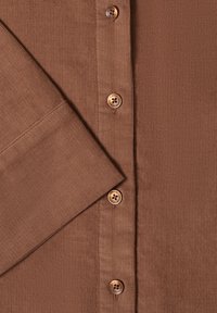 Tissu côtelé marron avec des boutons marron, une manche pliée et un détail de ligne de boutons sur un vêtement.