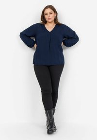 Marineblaues Langarmblusenoberteil mit V-Ausschnitt und subtilen Muster. Kombiniert mit schwarzen Skinny-Hosen und schwarzen Ankle-Boots. Glatte Textur, lockere Passform.