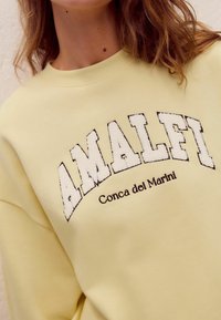 Gelbes Sweatshirt mit einem strukturierten, weißen gestickten "AMALFI"-Text und darunter in dunkler Fadenstickerei den kleineren "Conca dei Marini". Weicher Stoff.