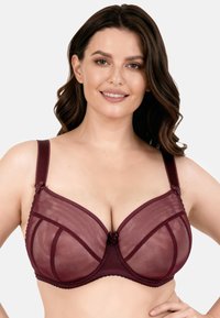 Usměvavá žena s vlnitými hnědými vlasy, která má na sobě průsvitnou vínovou podprsenku s kosticemi, pózuje s jednou rukou na bocích na bílém pozadí.