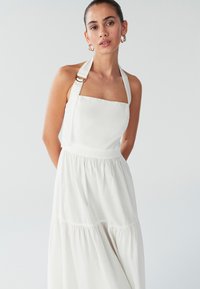 Vestido sin mangas de lino blanco con escote cuadrado, tirantes anchos con herrajes dorados y falda de volantes. Textura suave, ajuste casual.