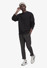 Juodas džemperis su užlenktais rankovėmis, tamsiai pilkos kelnės ir baltos sportinės bateliai su juodomis juostelėmis; modelis dėvi smėlio spalvos megztą skrybėlę.