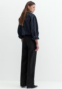 Kobieta ubrana w czarną, krótką kurtkę jeansową z podwiniętymi rękawami i czarne, szerokie spodnie z wysokim stanem, stojąca na białym tle.