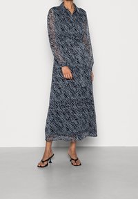 Robe longue bleu marine avec imprimé zèbre, manches longues transparentes, devant boutonné, taille ceinturée et silhouette fluide. Portée avec des sandales noires.