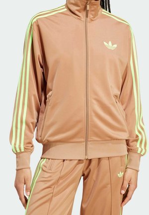 Person iført lysebrun træningsjakke og bukser med neongrønne striber og Adidas-logo på brystet og låret.