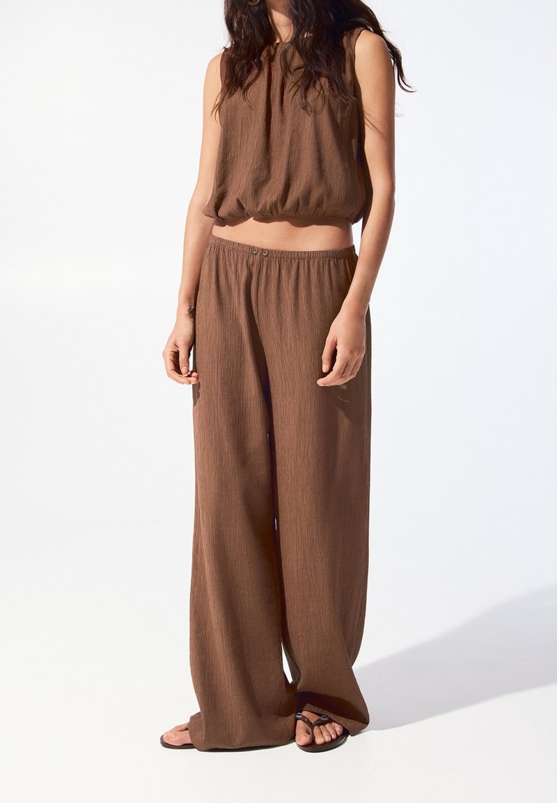 Jeune personne portant un haut ample marron sans manches et un pantalon large, debout pieds nus sur fond blanc.