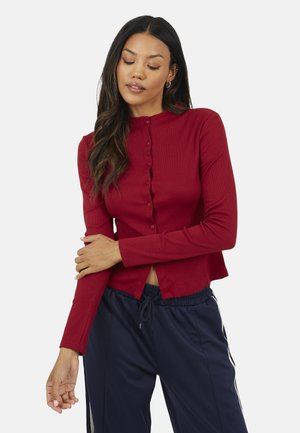 Donna con capelli lunghi e scuri che indossa una maglia rossa a maniche lunghe con bottoni e pantaloni blu navy con coulisse, in piedi contro uno sfondo chiaro.