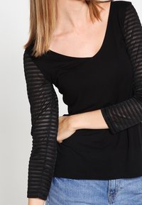 Schwarzes Oberteil mit tiefem V-Ausschnitt, ausgestattet mit gestreiften, durchsichtigen langen Ärmeln. Der Körper besteht aus glattem Stoff, während die Ärmel einen strukturierten Kontrast bieten.