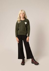 Olijfgroen gebreid sweater met een wit hartdetail, gecombineerd met zwarte wijde broek en bruine veterschoenen. Eenvoudig, textuurvol ontwerp.