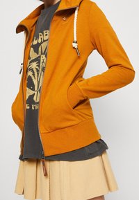 Giacca arancione con zip e cappuccio, caratterizzata da lacci a corda, indossata sopra una t-shirt grafica scura e una gonna in velluto a coste chiara.