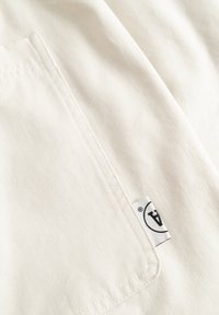 Gros plan sur un tissu écru avec une poche cousue et une petite étiquette blanche affichant un "A" noir à l'intérieur d'un cercle.