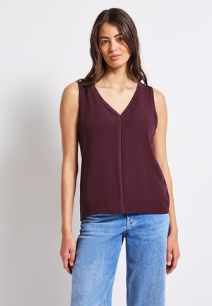Top bordeaux senza maniche con scollo a V, caratterizzato da delicate cuciture lungo il collo. Indossato con jeans larghi blu chiaro.