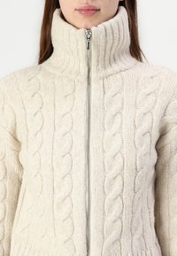 Sweater creme tricotado com gola alta e fecho total, apresentando um padrão texturizado e material suave e espesso.