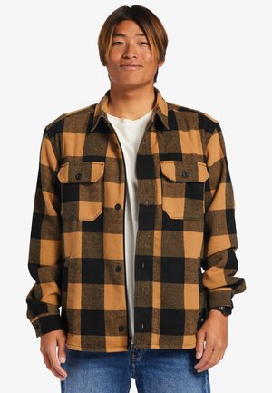 Brown and black plaid flannel shirt with two chest pockets, button-up front, and long sleeves, paired with a light-colored T-shirt and blue jeans.
