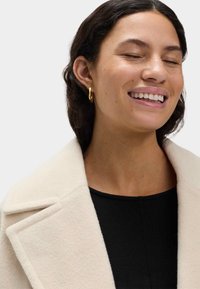 Cremefarbener Wollmischmantel mit Reverskragen und strukturierter Oberfläche. Modell trägt ein schwarzes Oberteil und zeigt kleine goldene Creolen.