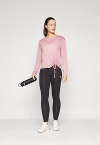 Light pink long-sleeve top with a gathered side detail, paired with black leggings. Holding a black water bottle, wearing white sneakers.