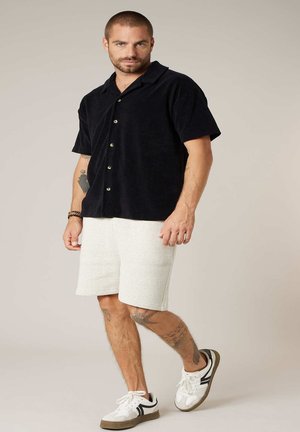 Homme aux cheveux courts et tatouages portant une chemise noire à manches courtes avec boutons, un short blanc texturé et des baskets blanches à rayures noires.