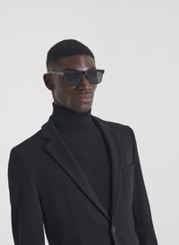 Blazer nero con rever a punta e taschino singolo, sovrapposto a un maglione a collo alto nero texturizzato. Indossato con occhiali da sole scuri e angolari.