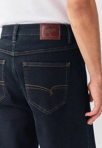 Tæt på en person iført mørkeblå denim jeans med orange syninger og et brunt lædermærke på taljen.