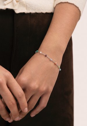 Muñeca con delicada pulsera de cadena de plata que presenta pequeñas cuentas redondas multicolores, sobre pantalones oscuros y puño de suéter beige claro.