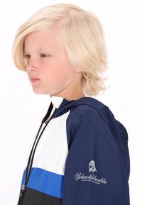 Marineblå og hvid hættejakke med en blå stribe. Har et logo på ærmet og et glat, letvægtsmateriale.