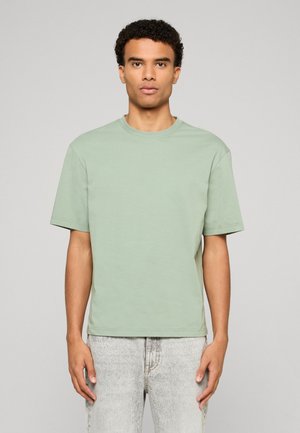 Jeune homme aux cheveux courts et bouclés, portant un t-shirt vert clair à manches courtes et un jean gris clair, debout devant un fond uni.