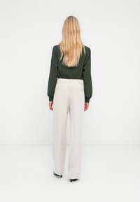 Blouse verte à manches longues avec épaules bouffantes associée à un pantalon large beige clair, mettant en valeur une texture lisse et des détails subtils.