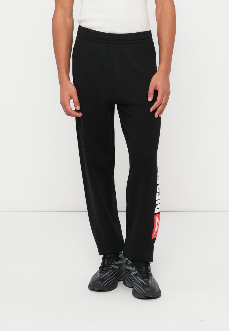 Μαύρο παντελόνι φόρμας με άνετη εφαρμογή, ελαστική μέση και λευκή/κόκκινη σήμανση στην αριστερή πλευρά. Το υλικό φαίνεται λείο με ελαφριά γυαλάδα.