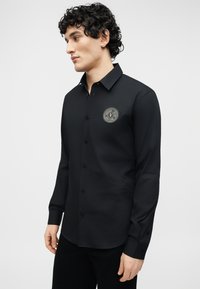 Άνδρας με σγουρά μαλλιά που φοράει μαύρο πουκάμισο με κουμπιά και λογότυπο Versace Jeans Couture στο αριστερό μέρος του στήθους, στέκεται μπροστά σε ένα απλό φωτεινό φόντο.