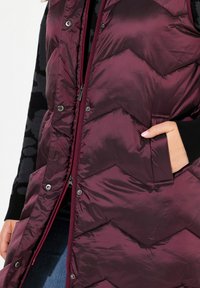 Gilet imbottito bordeaux con motivo a zigzag trapuntato, dotato di chiusura zip frontale, bottoni a pressione e tasche laterali. Tessuto liscio e lucido.