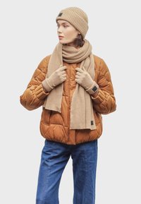 Bruine pufferjack, beige ribbelgebreide muts, sjaal en handschoenen. Casual look met lichtblauwe denim broek. Zachte texturen, warme kleuren.