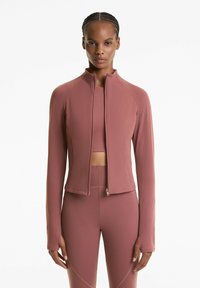 Žena oblečená v zladenom fialovom zip-up saku, crop top a legínach s vysokým pásom, stojaca na bielom pozadí.