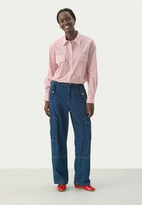 Camisa a rayas roja y blanca de botones con dos bolsillos frontales, combinada con pantalones de carga de mezclilla azul con bolsillos, usados con flats rojos.