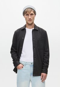 Σκούρο γκρι πουκάμισο με γιακά, μία θήκη για το στήθος και μαύρα κουμπιά, φορεμένο πάνω από λευκό t-shirt, συνδυασμένο με ανοιχτό μπλε τζιν.