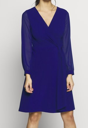 Vrouw die een koningsblauwe wikkeljurk tot op de knie draagt met doorschijnende lange mouwen en een V-hals, staande tegen een effen lichte achtergrond.
