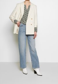 Blazer crème croisé, blouse noire et blanche à motifs, jeans large bleu clair et bottines blanches à talons.