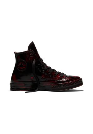 Zapatilla negra de caña alta con acabado brillante, cordones negros y un parche redondo con logo en el tobillo, vista lateral sobre fondo blanco.