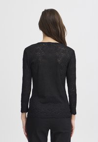 Top in pizzo nero con maniche lunghe, caratterizzato da un motivo a spirale e orlo scallop. Texture semi-trasparente che rivela la pelle sottostante.