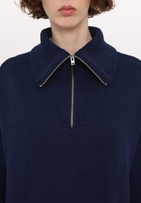 Gros plan d'une personne portant un pull en maille bleu marine avec une fermeture éclair argentée demi-zip et un large col plié.