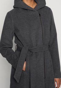 Abrigo gris con capucha y cintura con cinturón, con cremallera de longitud completa y bolsillos laterales. Hecho de tela suave y texturizada.