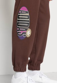 Hnědé tepláky s velkým vícebarevným logem Adidas na boku, s žebrovaným manžetovým lemem na kotníku, nošené s bílými teniskami.