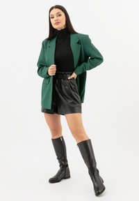 Blazer verde, dolcevita nero, pantaloncini in pelle nera e stivali neri al ginocchio. Caratterizzato da linee eleganti e una vestibilità comoda.
