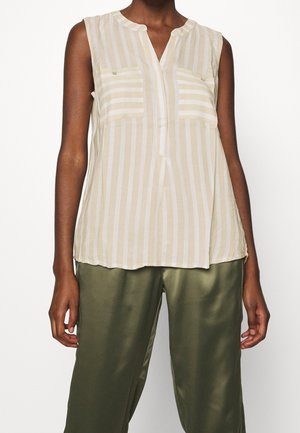 Le modèle porte un chemisier crème rayé sans manches, à col en V et à poches poitrine, associé à un pantalon vert olive.