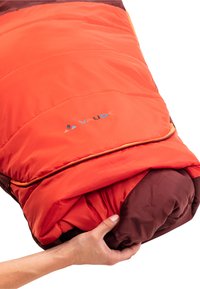 Sac de couchage rouge et brun, avec un design matelassé et une fermeture éclair visible. Main tenant le sac roulé. Logo imprimé sur la surface.