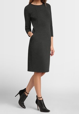 Femme portant une robe gris foncé longueur genoux avec des manches trois-quarts et des bottines noires pointues à talons hauts avec des brides à la cheville.