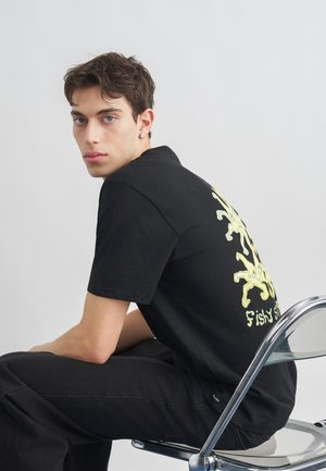 Νεαρός άνδρας με σκοτεινά μαλλιά κάθεται πλαγίως σε διάφανη καρέκλα, φορόντας μαύρο μπλουζάκι με κίτρινες σχεδιάσεις και μαύρο παντελόνι, με φόντο ένα απλό σκηνικό.