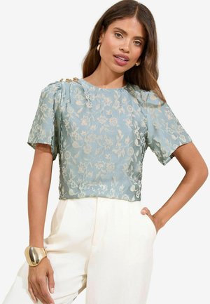 Light blue short-sleeve blouse with floral lace pattern and pleated shoulders. Accentuated by gold hardware on the shoulder. Worn with white trousers.
