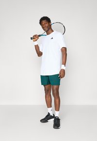 T-shirt de sport blanc avec logo noir, shorts verts, bandeaux blancs aux poignets et chaussures de tennis noires. Tenant une raquette de tennis, se tenant debout devant un fond neutre.